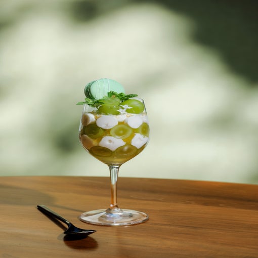 Glass dish with green grapes, plant-based cream, and a green macaron topped with mint; placed on a wooden table next to a dark spoon.