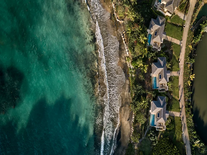 Seaside villas with private pools, surrounded by greenery, adjacent to a sandy shore where gentle waves meet the clear, turquoise ocean. Accessible paths everywhere.