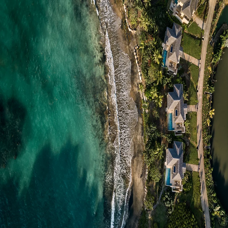 Seaside villas with private pools, surrounded by greenery, adjacent to a sandy shore where gentle waves meet the clear, turquoise ocean. Accessible paths everywhere.