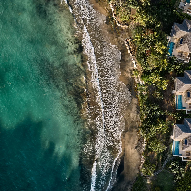 Seaside villas with private pools, surrounded by greenery, adjacent to a sandy shore where gentle waves meet the clear, turquoise ocean. Accessible paths everywhere.
