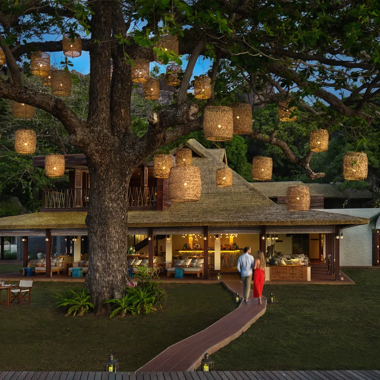 Guests approach a softly illuminated villa at dusk, with lanterns lighting the path beneath a large tree, reflecting a tranquil atmosphere.