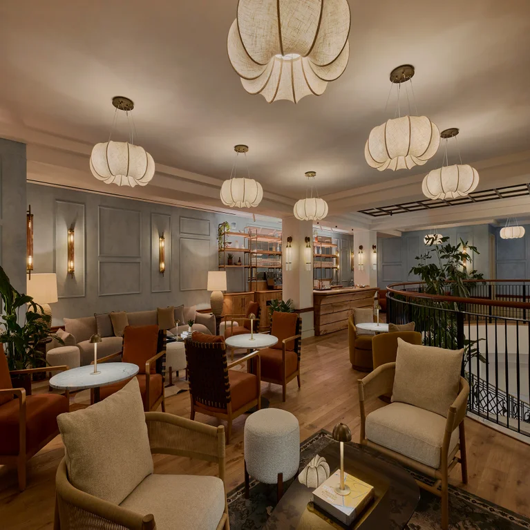 Lounge space featuring neutral-tone armchairs, circular tables, gentle lighting, pendant ceiling lights, and a wall of bookshelves.