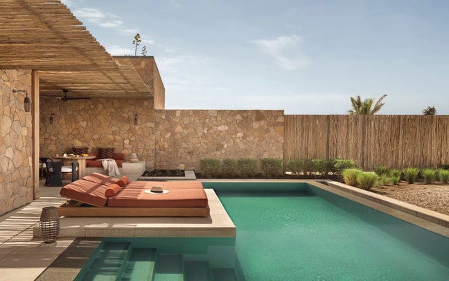 Poolside with two cushioned lounge chairs, a straw hat resting nearby, natural stone walls, wooden pergola overhead, and lush plants beneath an open sky.