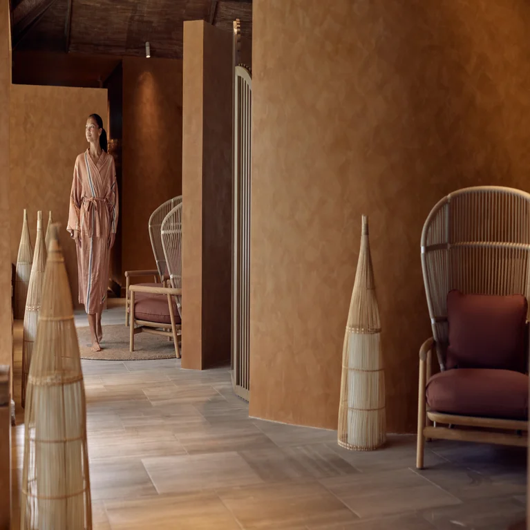Guest in a robe moves through a softly lit spa lounge, designed with natural wicker seating and woven decor accents.