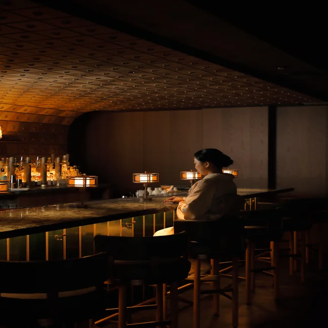 Person in cultural dress sits at a softly illuminated bar with wood finishes and lantern lights, evoking warmth and inviting ambiance.