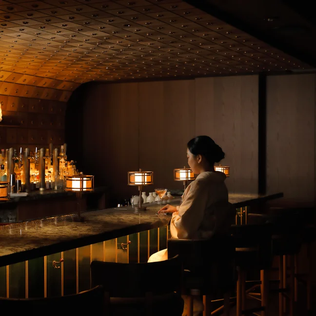 Person in cultural dress sits at a softly illuminated bar with wood finishes and lantern lights, evoking warmth and inviting ambiance.