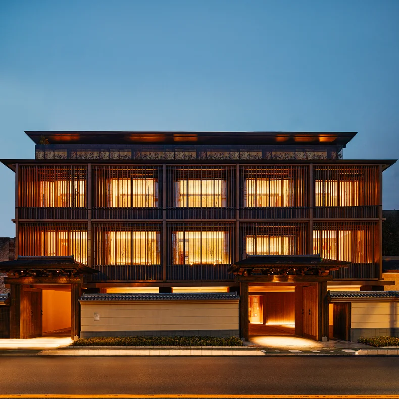 Building inspired by Japanese design, featuring wooden slats and illuminated windows along the facade at dusk, with a focus on sustainable architecture.