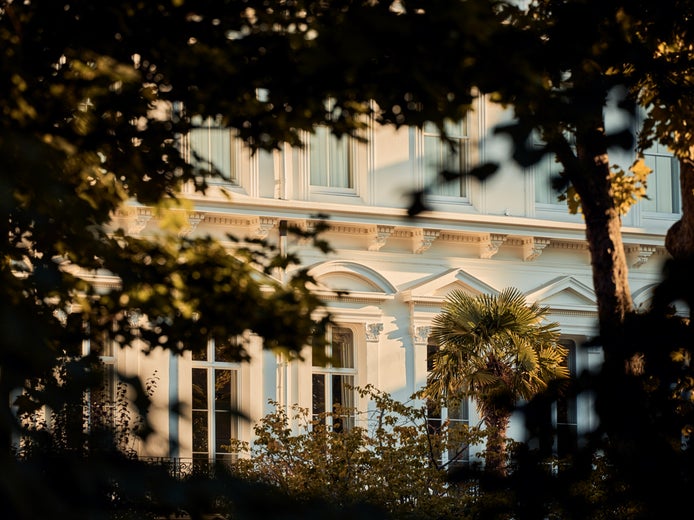 Một tòa nhà màu trắng với những ô cửa sổ rộng lớn, được bao quanh bởi những cây cổ thụ và ngập tràn ánh sáng tự nhiên, thể hiện sự hài hòa giữa thiết kế hiện đại và sự hòa hợp với thiên nhiên.