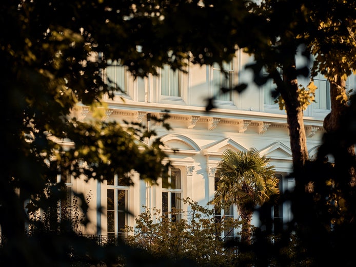 Một tòa nhà màu trắng với những ô cửa sổ rộng lớn, được bao quanh bởi những cây cổ thụ và ngập tràn ánh sáng tự nhiên, thể hiện sự hài hòa giữa thiết kế hiện đại và sự hòa hợp với thiên nhiên.