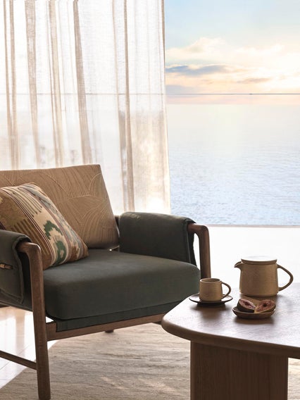 An armchair with cushions is positioned near a window facing the sea, alongside a table set with a teapot and cups in gentle daylight.