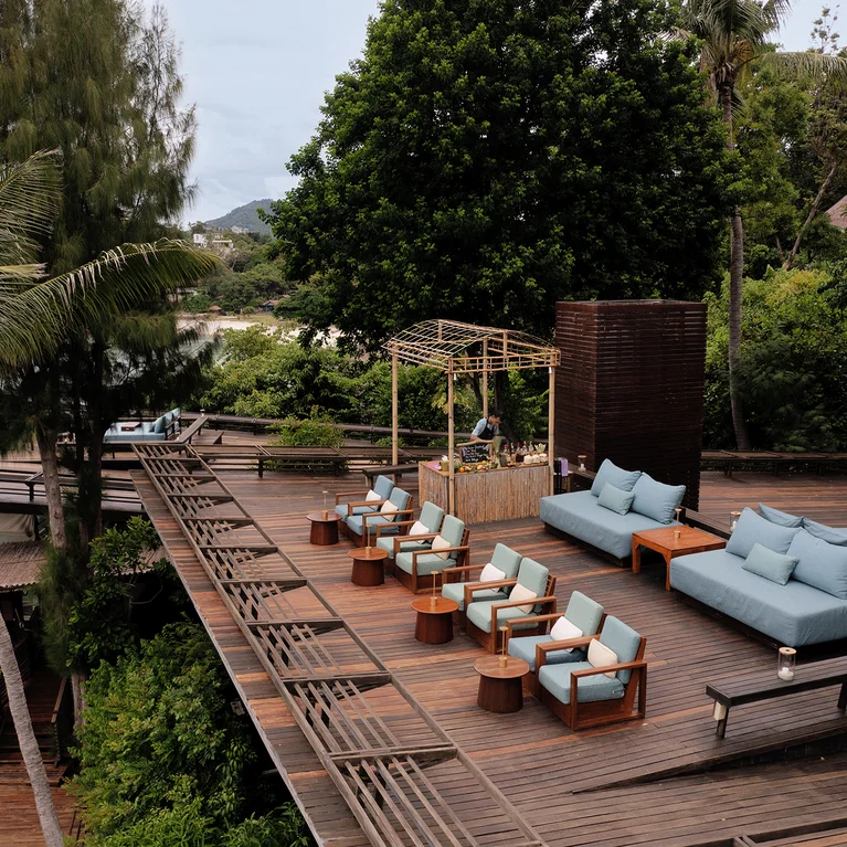 木质甲板上摆放着蓝色沙发、扶手椅和一个小酒吧,棕榈树和绿树掩映在平静的水面上。小径宽阔,便于通行。.