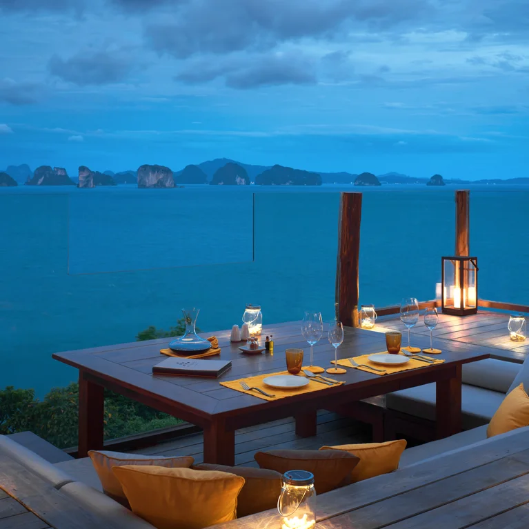 Une table en bois pour quatre personnes sur une terrasse avec des lanternes et des sièges rembourrés, face à l'océan au coucher du soleil avec des îles lointaines sous un ciel nuageux.