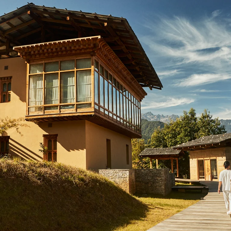 Un huésped pasea por un sendero de madera entre edificios tradicionales con tejados de madera, con cielos azules, nubes blancas y montañas lejanas....