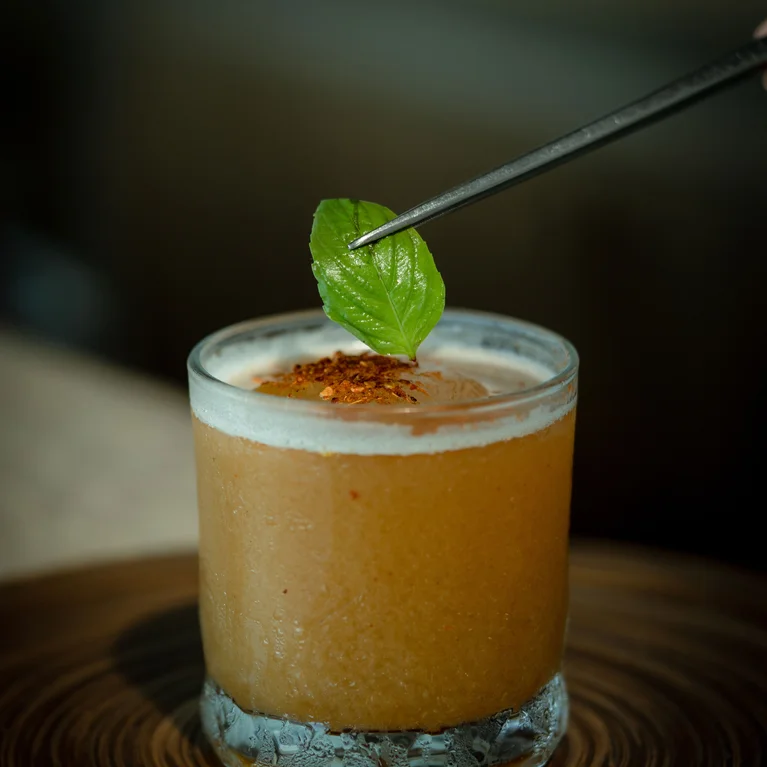 يوضع الملقط برفق ورقة خضراء طازجة فوق كوكتيل بني اللون برغوة تُقدم في كوب قصير على صينية خشبية مستديرة.