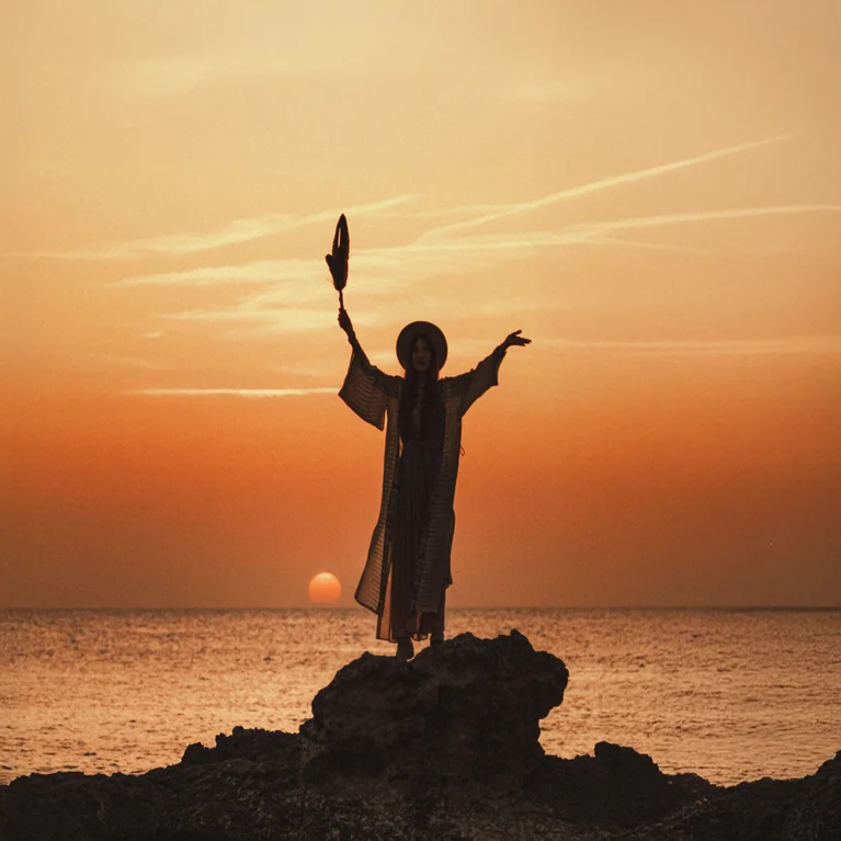 Individual wearing a robe and hat stands on coastal rocks at sunset, holding an item aloft with arms raised toward the orange sky over the sea.