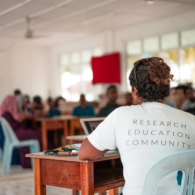 An individual in a Research Education Community t-shirt sits at a desk, engaging with others in an open, light-filled learning space.