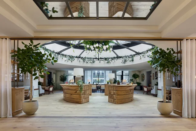 Lobby featuring natural wood desks, abundant indoor plants, and a skylight ceiling with suspended greenery and orb lighting, emphasizing wellness and biophilic design.