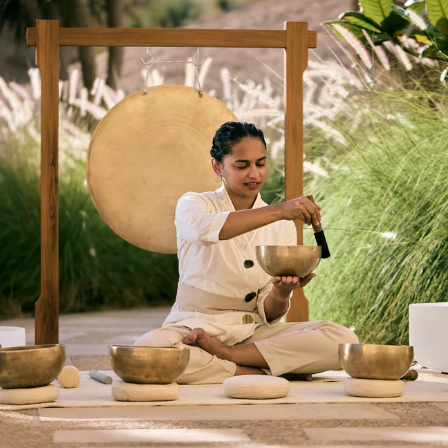 An individual plays singing bowls with a mallet in an outdoor setting. A large gong and abundant green plants create a calming, natural atmosphere.