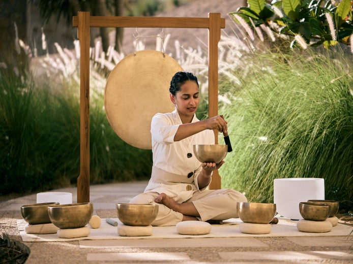 An individual plays singing bowls with a mallet in an outdoor setting. A large gong and abundant green plants create a calming, natural atmosphere.
