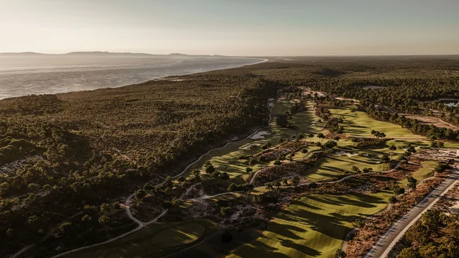 Golf course bordered by lush trees, with ocean coastline on the left and gentle hills visible in the distance under a clear sky.