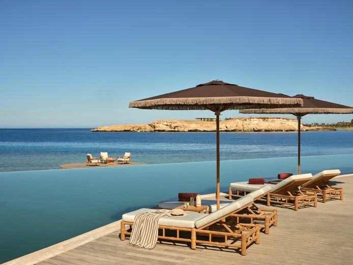 Lounge chairs with umbrellas beside an infinity pool face a tranquil sea; a table with chairs is set on a sandbar under a clear sky in the distance.