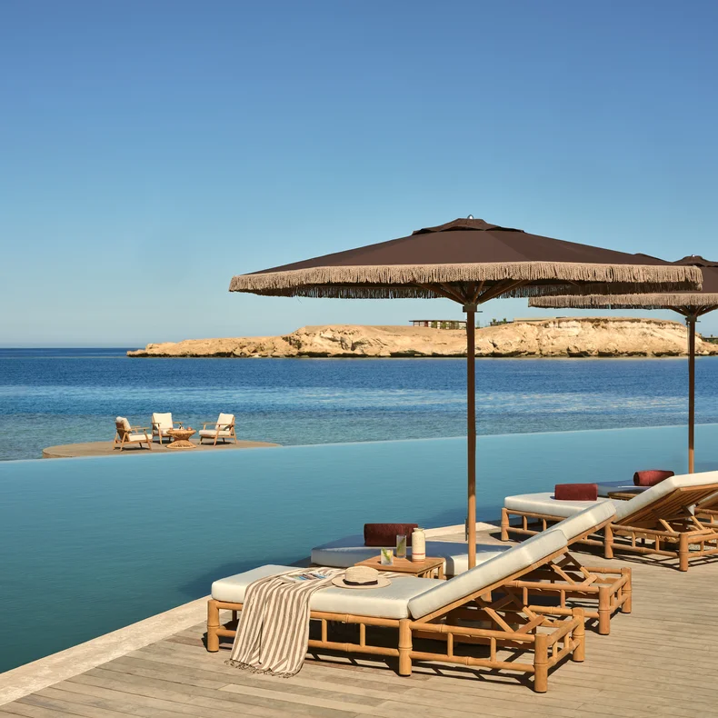 Lounge chairs with umbrellas beside an infinity pool face a tranquil sea; a table with chairs is set on a sandbar under a clear sky in the distance.