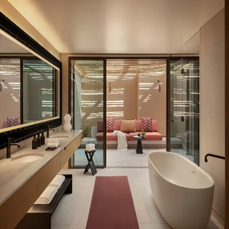 Serene bathroom featuring a deep soaking tub, twin sinks, generous mirror, walk-in glass shower, and doors leading to a patio with outdoor seating and cushions.