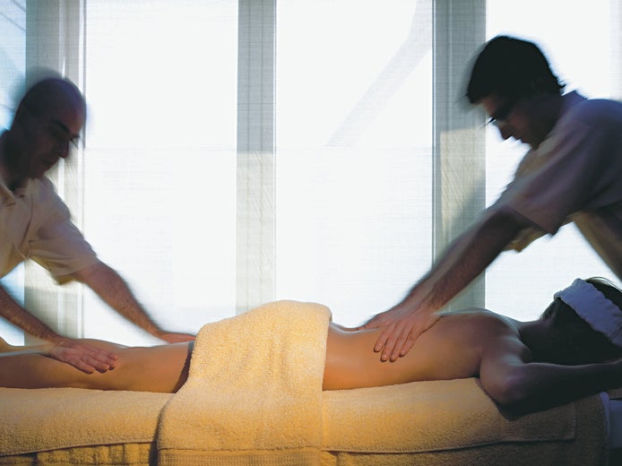 Therapists provide massage for a guest lying face down on a table, draped with towels, in a room gently illuminated for comfort and relaxation.