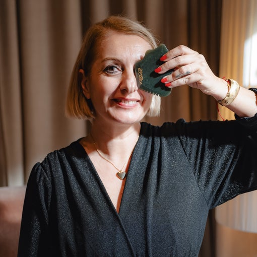 A guest gently uses a green gua sha stone on her face in a softly lit space, with natural brown curtains creating a calm, inviting atmosphere.