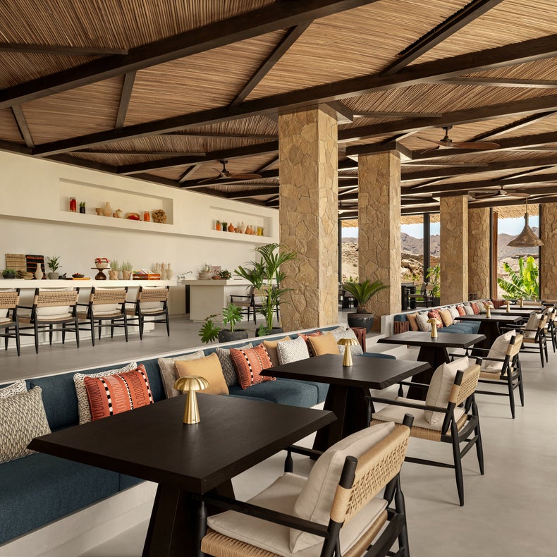 Open-air dining area featuring stone columns, a wooden ceiling, tables, plush seating, and shelves. Ample daylight with mountain vistas supports calm, mindful relaxation.