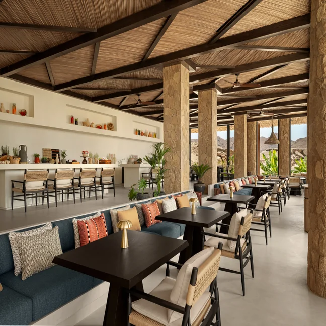 Open-air dining area featuring stone columns, a wooden ceiling, tables, plush seating, and shelves. Ample daylight with mountain vistas supports calm, mindful relaxation.