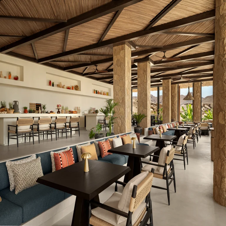 Open-air dining area featuring stone columns, a wooden ceiling, tables, plush seating, and shelves. Ample daylight with mountain vistas supports calm, mindful relaxation.