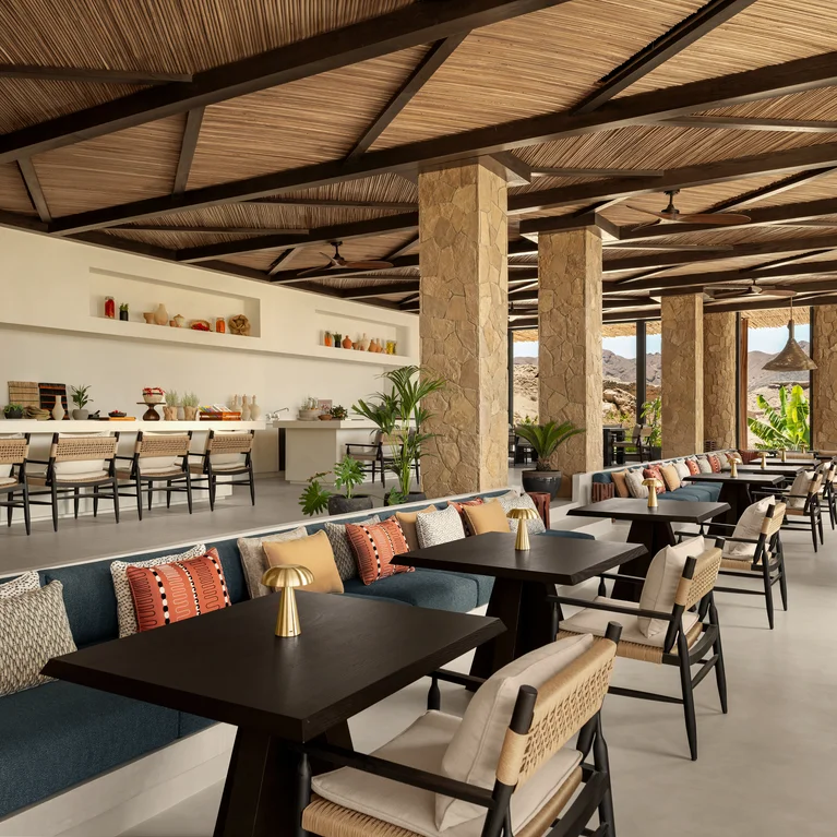 Open-air dining area featuring stone columns, a wooden ceiling, tables, plush seating, and shelves. Ample daylight with mountain vistas supports calm, mindful relaxation.