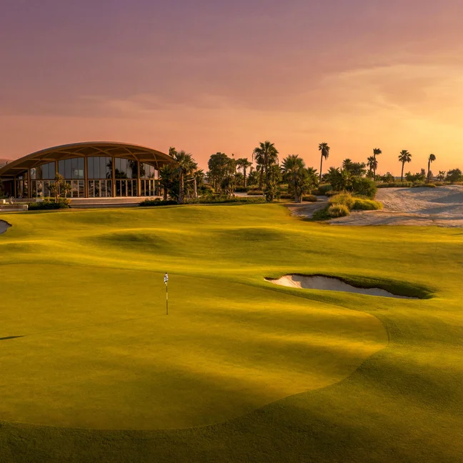 Golf course featuring flagstick, sand bunkers, palm trees, and a contemporary clubhouse under warm evening light. Accessible pathways are visible throughout.