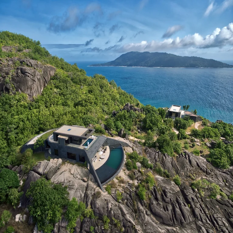 Villa with pool on a rocky hillside, surrounded by greenery, overlooking ocean and a distant island under a partly cloudy sky. Sustainable design focus.