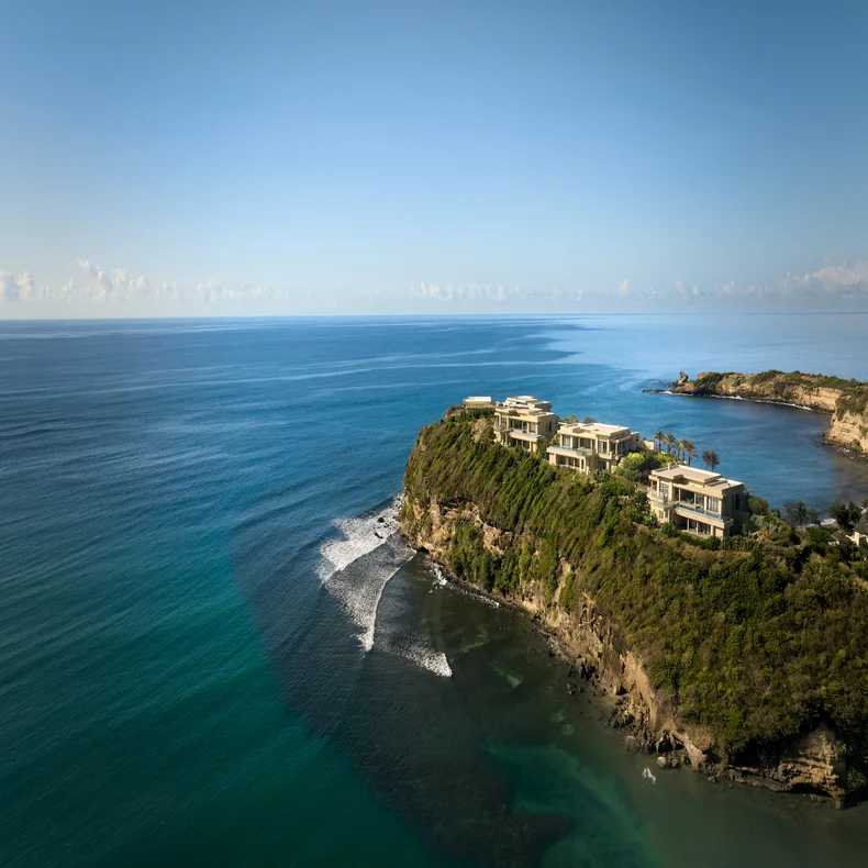Los edificios contemporáneos construidos sobre un acantilado verde se asoman al océano turquesa, con suaves olas batiendo la costa rocosa bajo un cielo azul despejado.