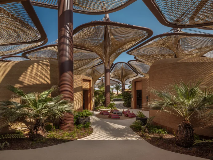 Spacious plaza features woven canopies, curved architecture, palm trees, and lounge seating, designed for comfort and shaded relaxation beneath a clear blue sky.