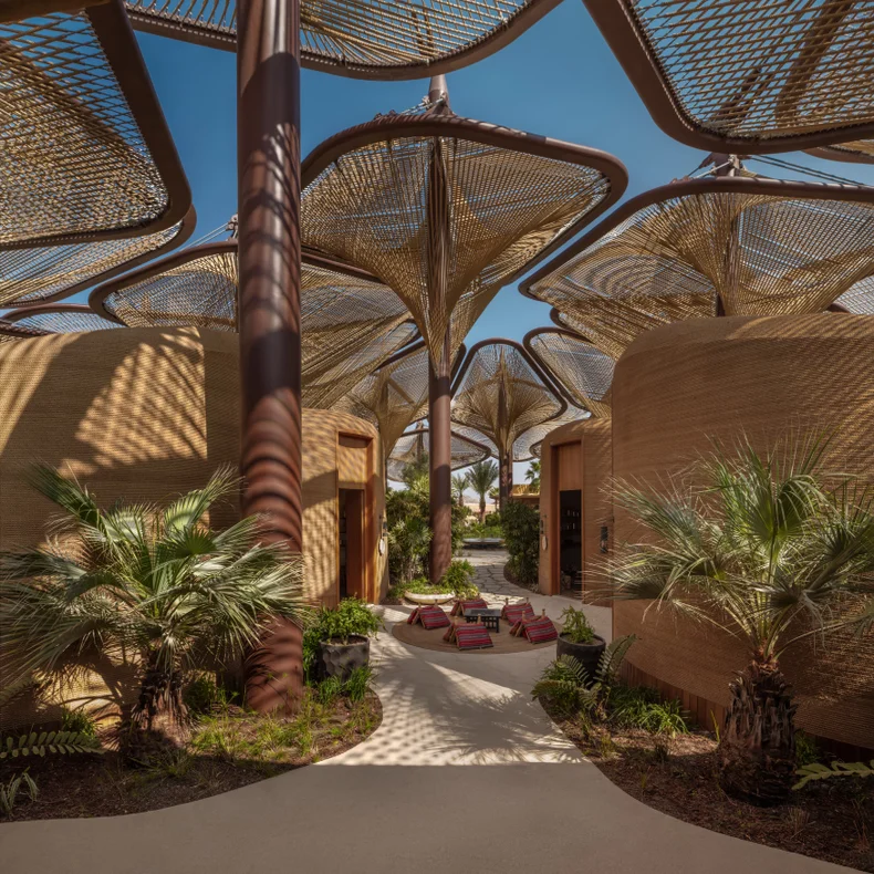 Spacious plaza features woven canopies, curved architecture, palm trees, and lounge seating, designed for comfort and shaded relaxation beneath a clear blue sky.