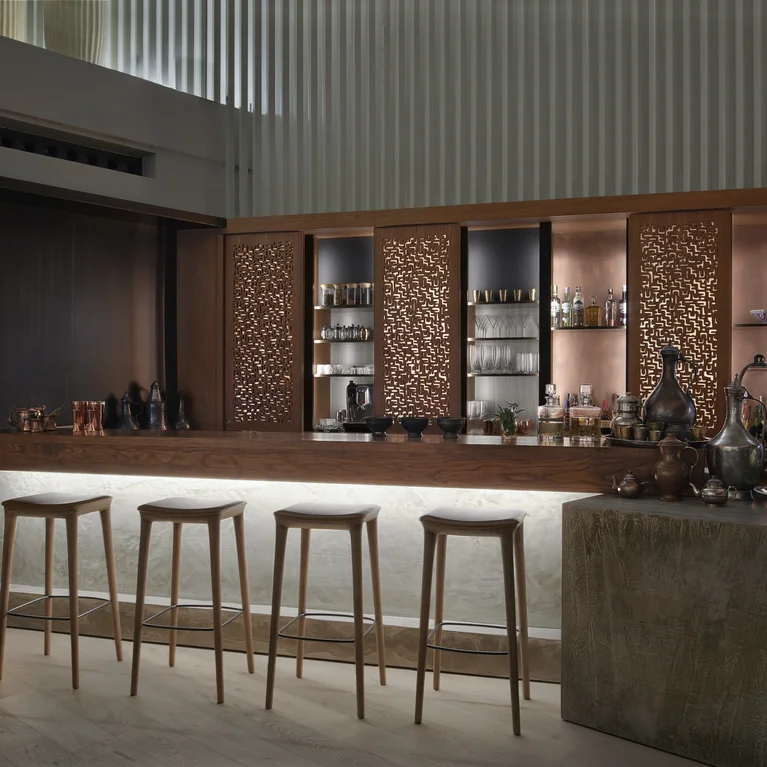 La zona del bar incluye cuatro taburetes de madera, un mostrador de piedra de hormigón, pantallas de madera tallada, estanterías organizadas con botellas y cristalería, y una suave iluminación difusa.