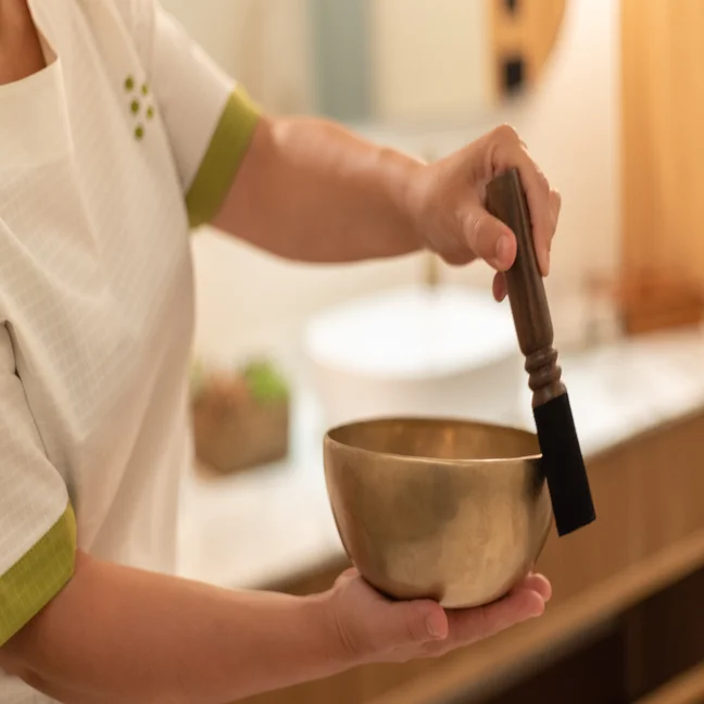 Hands gently hold a brass singing bowl and wooden mallet, ready for use in a softly lit, calming space designed for mindfulness.