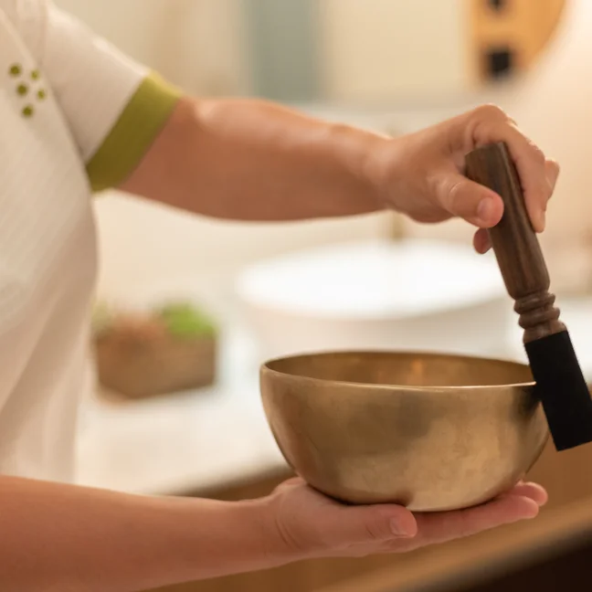 Hands gently hold a brass singing bowl and wooden mallet, ready for use in a softly lit, calming space designed for mindfulness.
