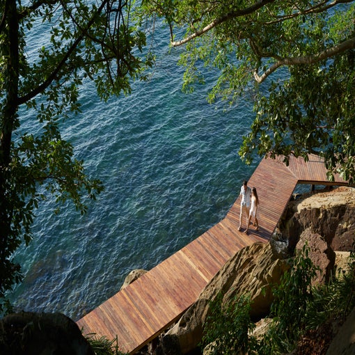 Guests stroll along a wooden boardwalk beside clear blue water, with trees and rocks nearby, as dappled sunlight filters through the leaves.