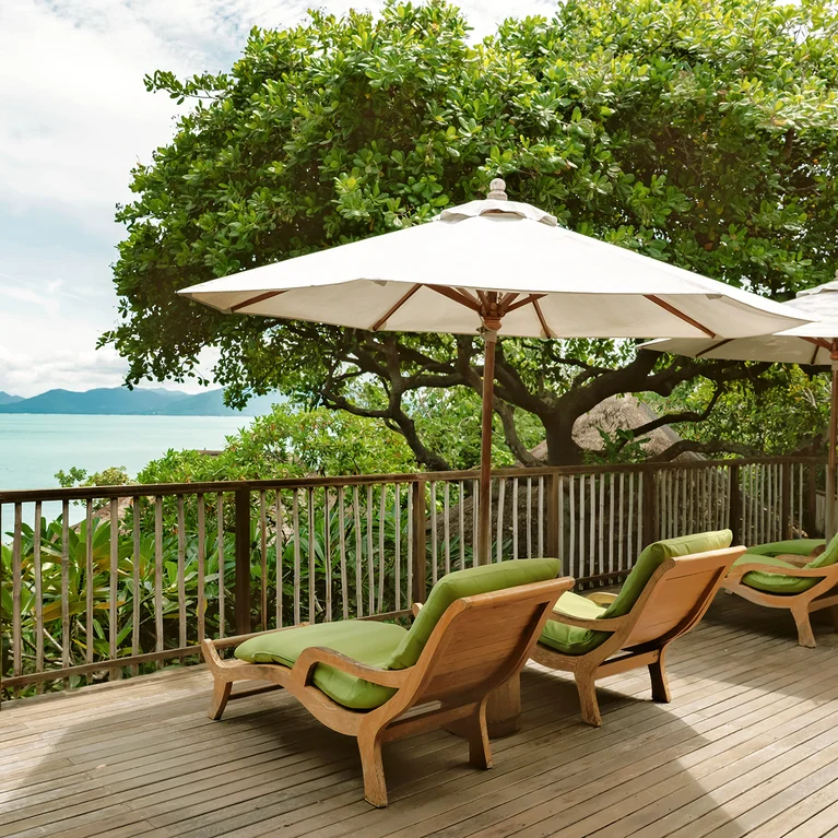تصطف كراسي التشمس ذات الوسائد الخضراء على سطح خشبي مظلّل بمظلات بيضاء تطل على المحيط والأشجار المورقة، مما يخلق منطقة استرخاء هادئة في الهواء الطلق.