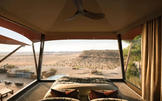 A serene bedroom with expansive glass windows opens to views of desert cliffs and native plants, with a private outdoor hot tub on a wooden deck.