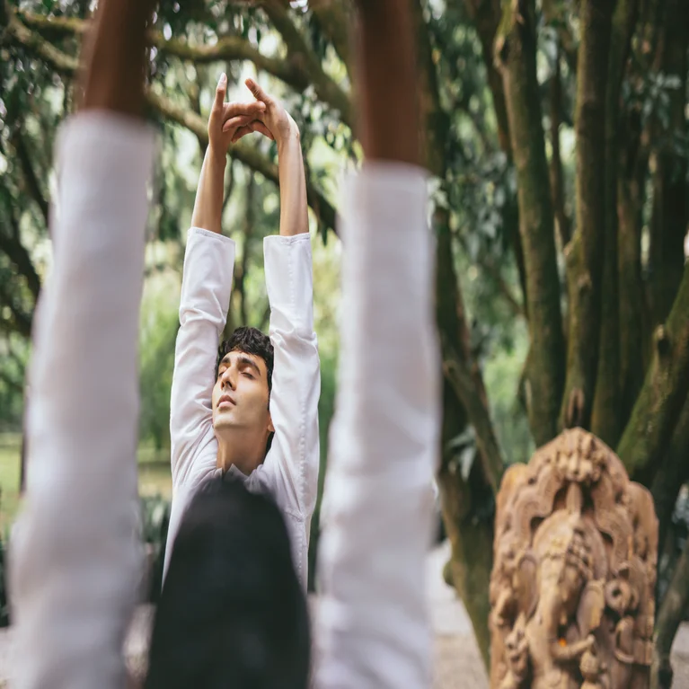A guest practices mindful movement outdoors beside a mature tree and detailed sculpture, with another guest softly out of focus in the foreground.