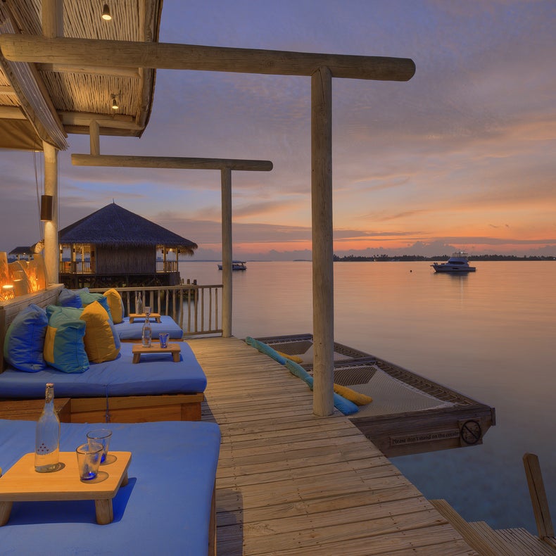 Serene seating area with blue cushions and vibrant pillows on a wooden deck above calm water at sunset, drinks on tables, boats visible in the distance.