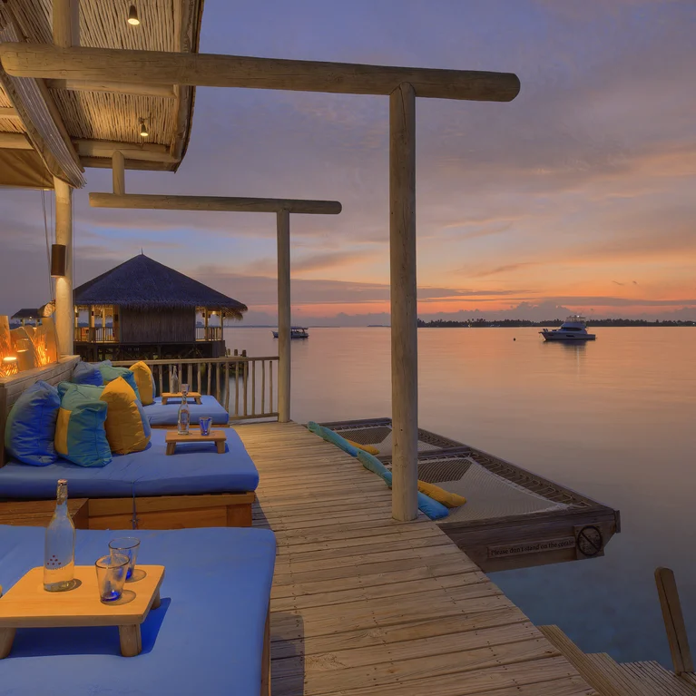 Serene seating area with blue cushions and vibrant pillows on a wooden deck above calm water at sunset, drinks on tables, boats visible in the distance.