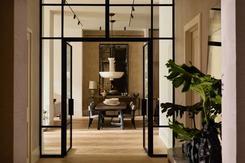 Dining room with glass double doors, wood table, dark chairs, a chandelier above, leafy plant nearby, and light herringbone flooring creating a serene atmosphere.