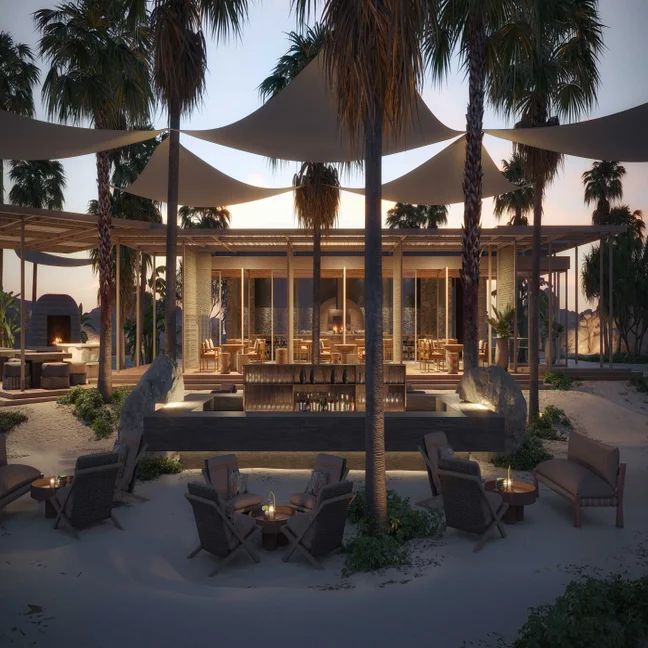Outdoor lounge space with seating, tables, palm trees, and fire pits on sand. Soft evening lights glow beneath expansive shade sails for relaxed beachfront gatherings.