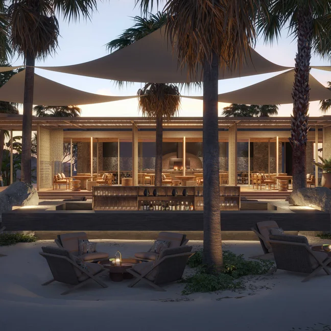 Outdoor lounge space with seating, tables, palm trees, and fire pits on sand. Soft evening lights glow beneath expansive shade sails for relaxed beachfront gatherings.